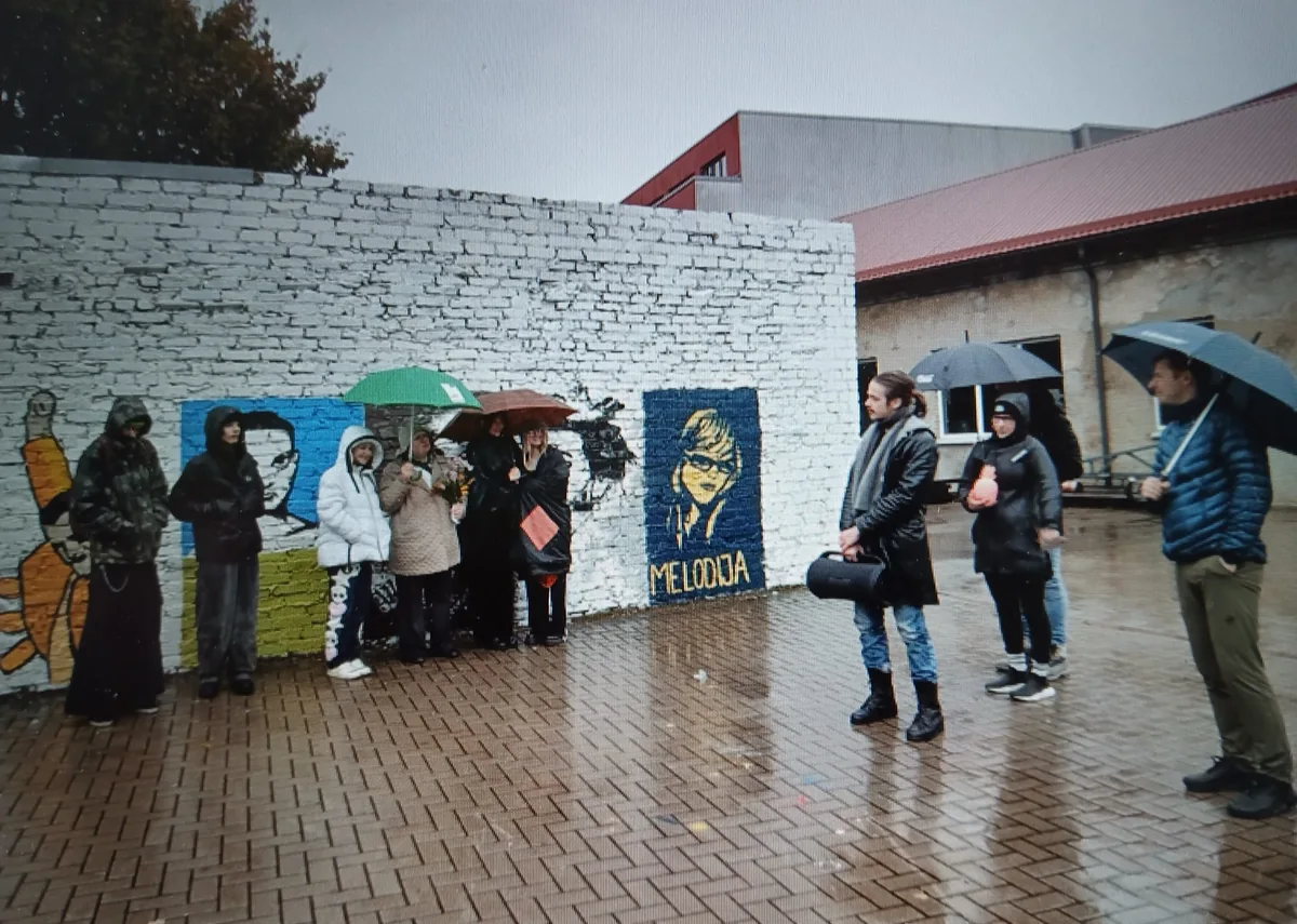 Cilvēki ar lietussargiem pie apgleznotas ķieģeļu sienas ar uzrakstu “MELODIJA”
